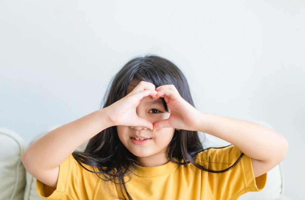 A young girl creates a heart shape with her hands and looks through it with her left eye.