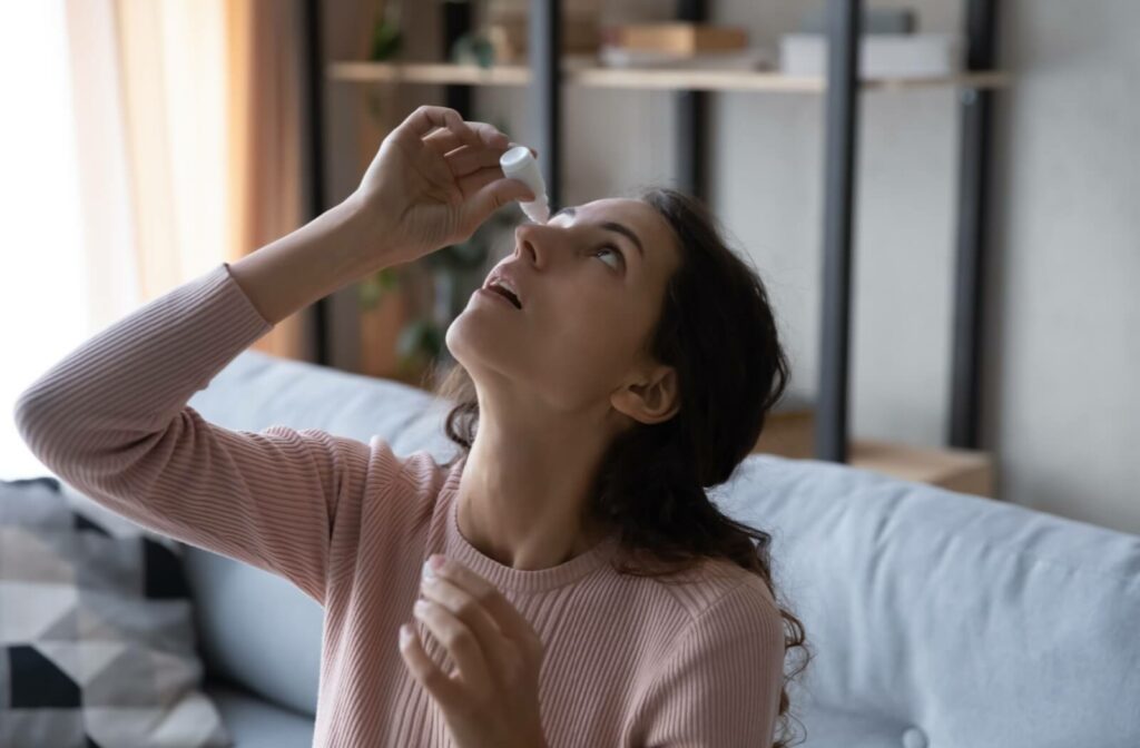 A person sits on their couching a brightly-lit living room, applying eye drops to one eye.