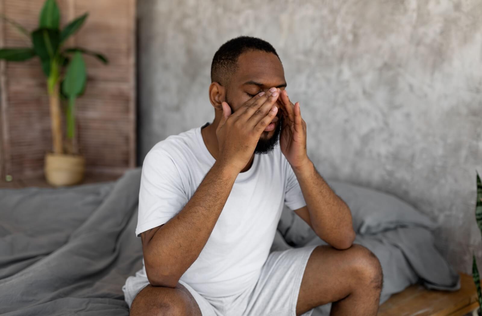 A person sits on the edge of his bed, just having woken up. He's resting his head on his hands and rubbing his eyes.