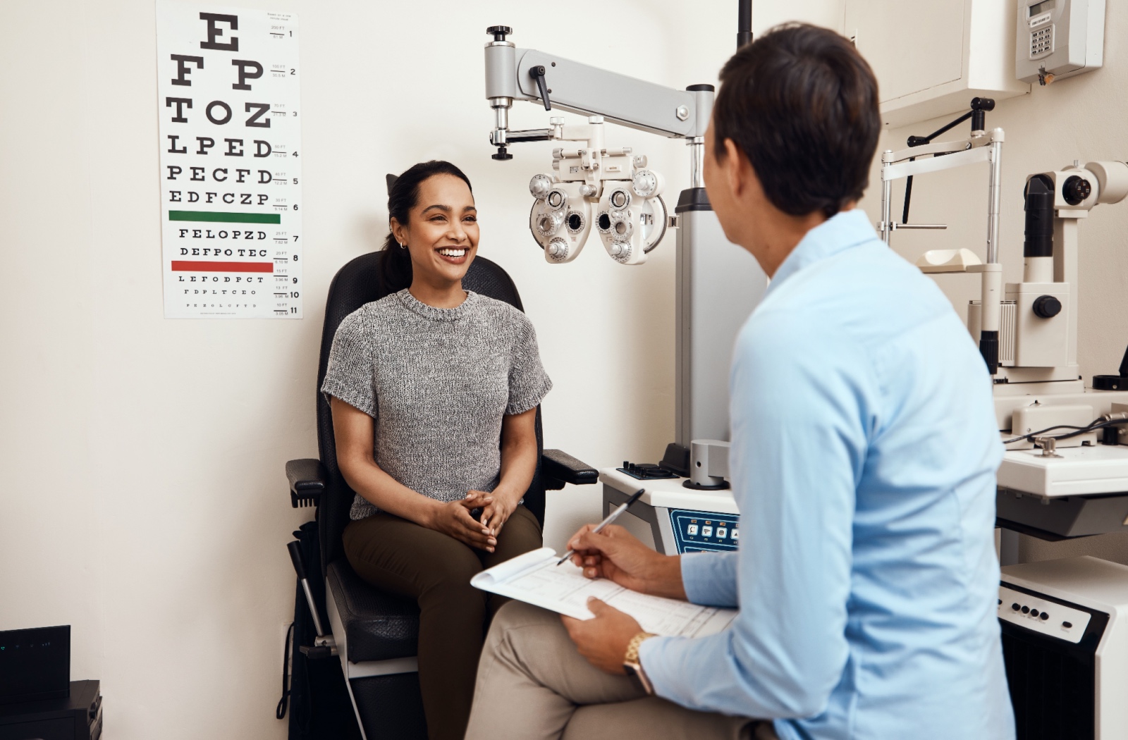 A smiling patient sits in an exam chair facing their optometrist, who is asking questions as part of an eye exam.