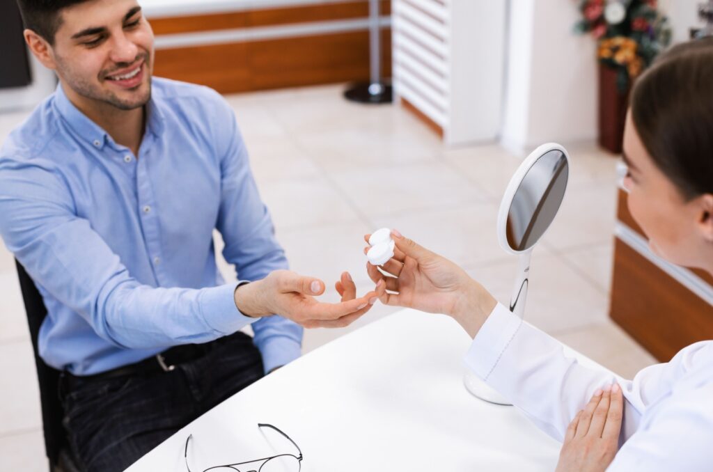 An eye doctor passing a contact lens case containing contact lenses to a smiling patient sitting at a table across from them.