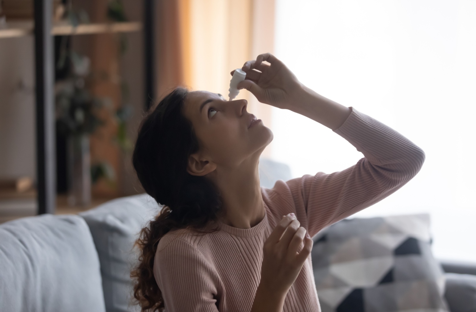 A young woman sitting on the couch putting in eye drops.