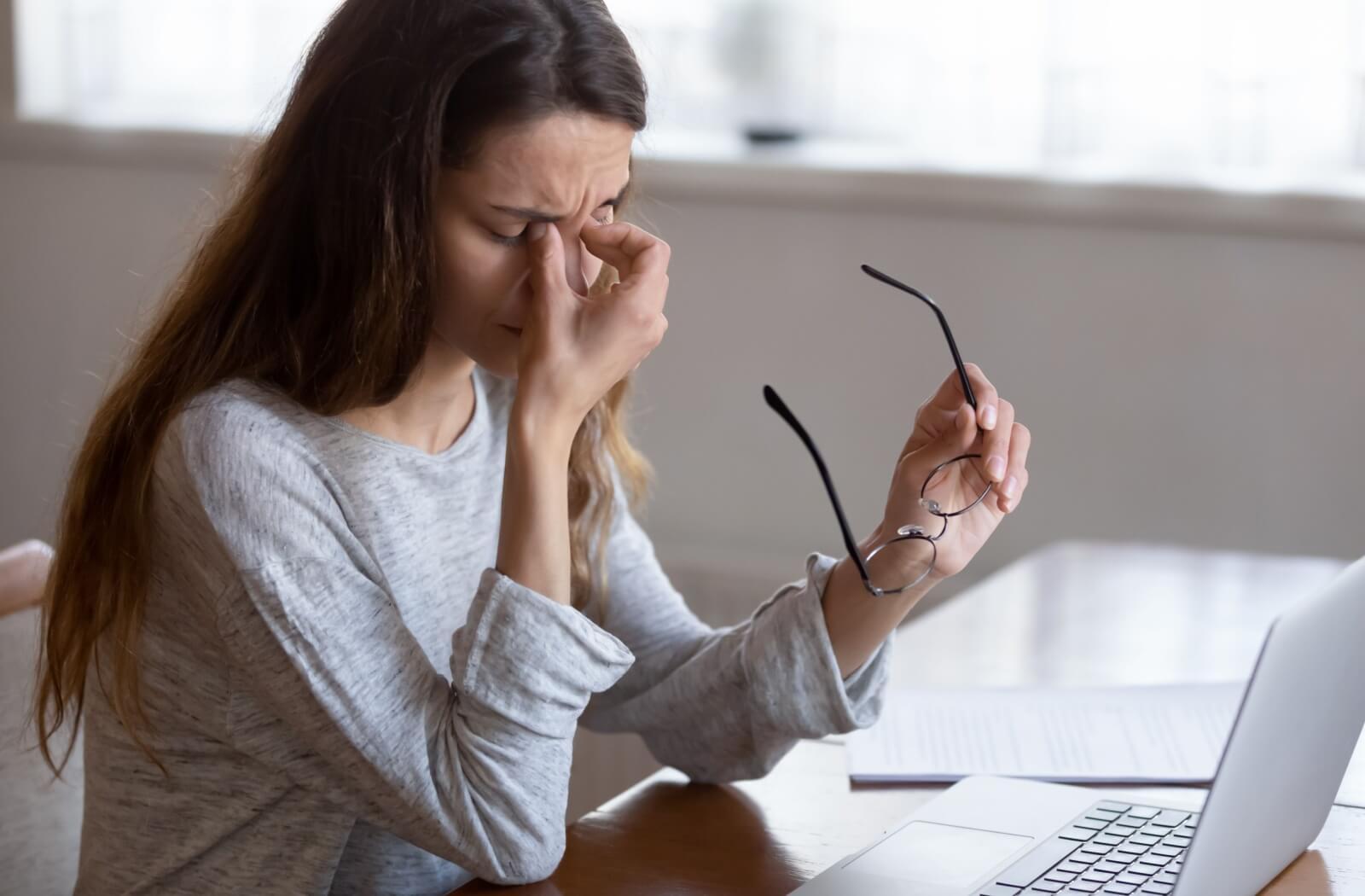 A young woman removing her glasses and pinching her nose as she takes a break from her computer screen.