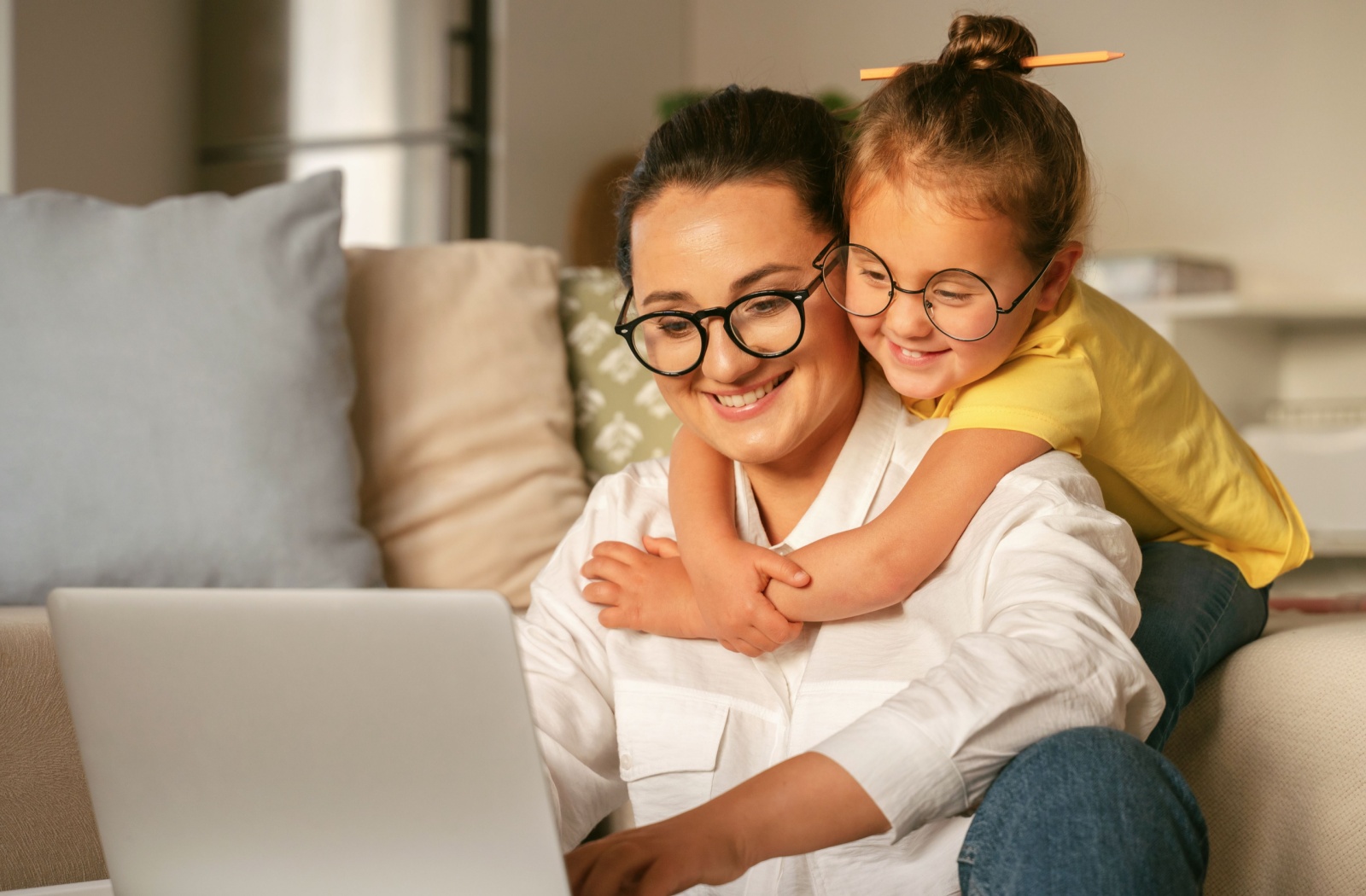 A child wearing round glasses hugs their parent, also wearing glasses, from behind while they work on a computer.